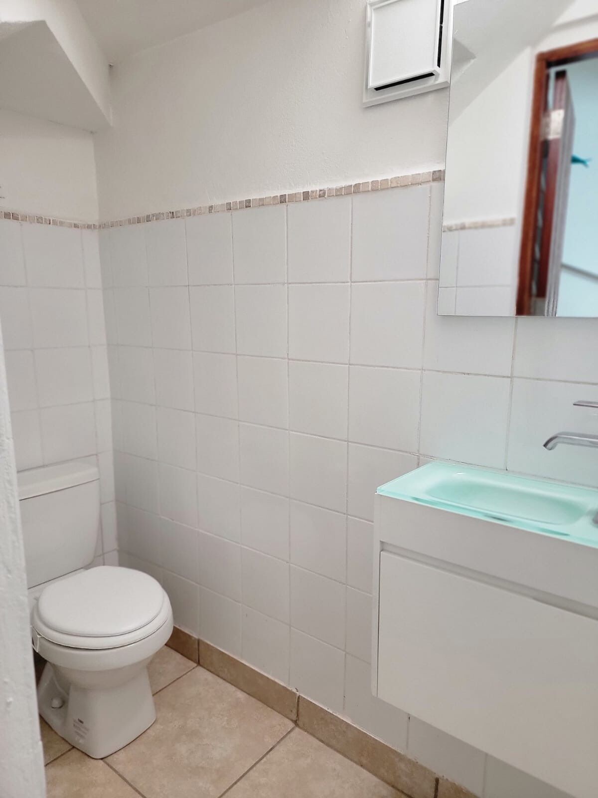 A small, clean bathroom with white tiles, a white toilet, and a light blue sink.