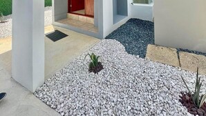 Un edificio moderno de dos plantas con diseño minimalista, que presenta un espacio central abierto con una puerta roja, una pequeña zona ajardinada con grava y plantas, y un cielo despejado.