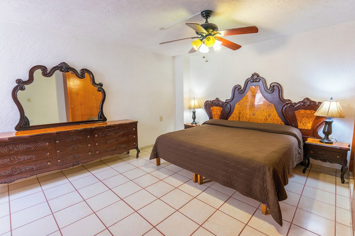 A bedroom with a bed, dresser, nightstand, lamp, ceiling fan, and a mirror.