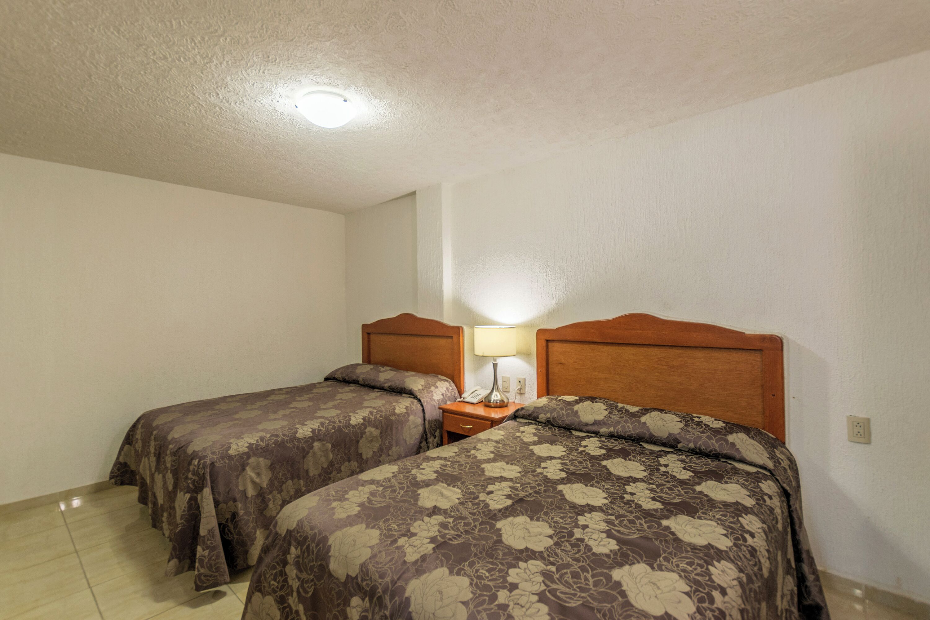 Quarto com duas camas, cada uma com cabeceira de madeira e colcha floral. Entre as camas, um criado-mudo com uma lâmpada e um telefone.