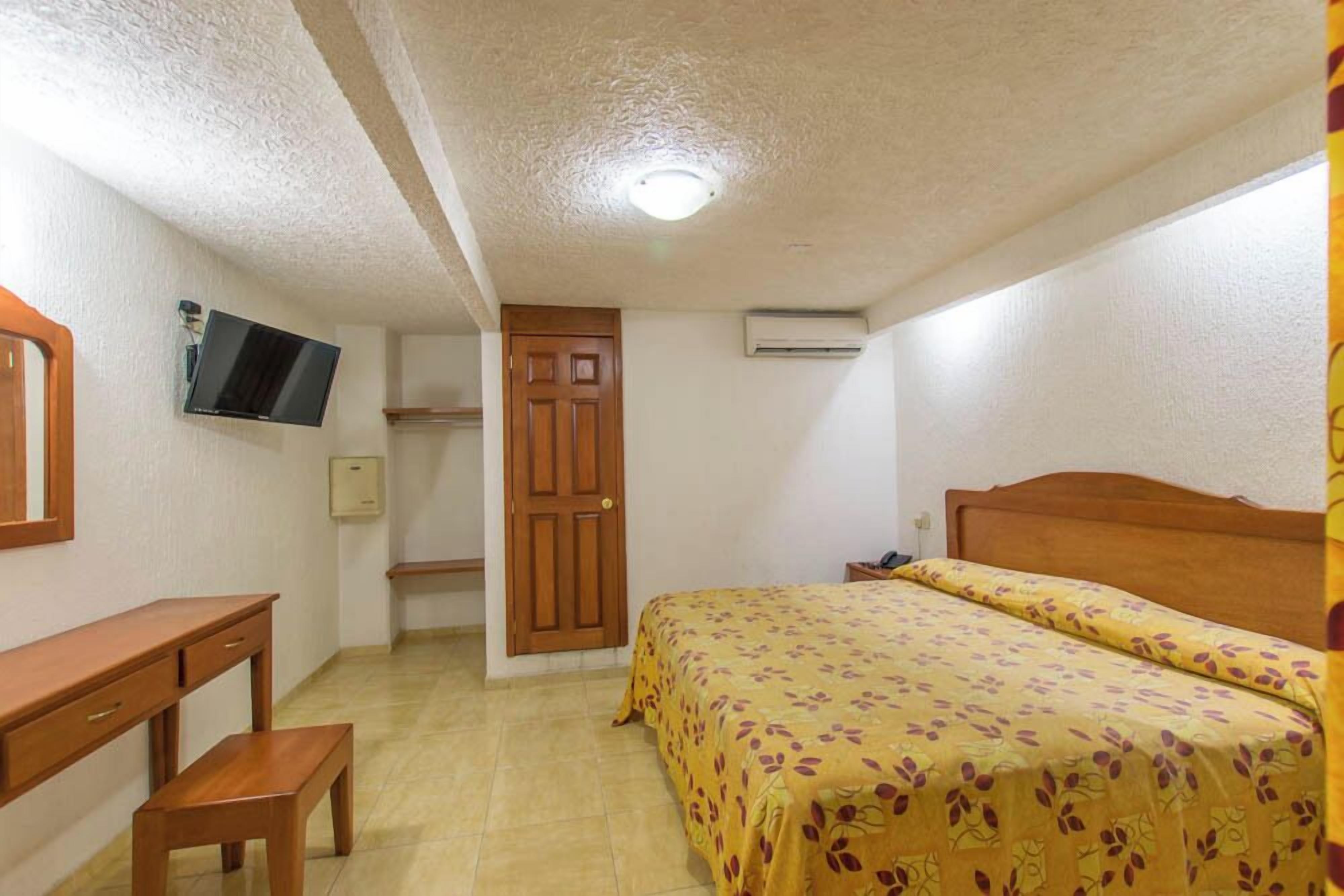 Un dormitorio con cama, escritorio, silla, televisor montado en la pared y una puerta.