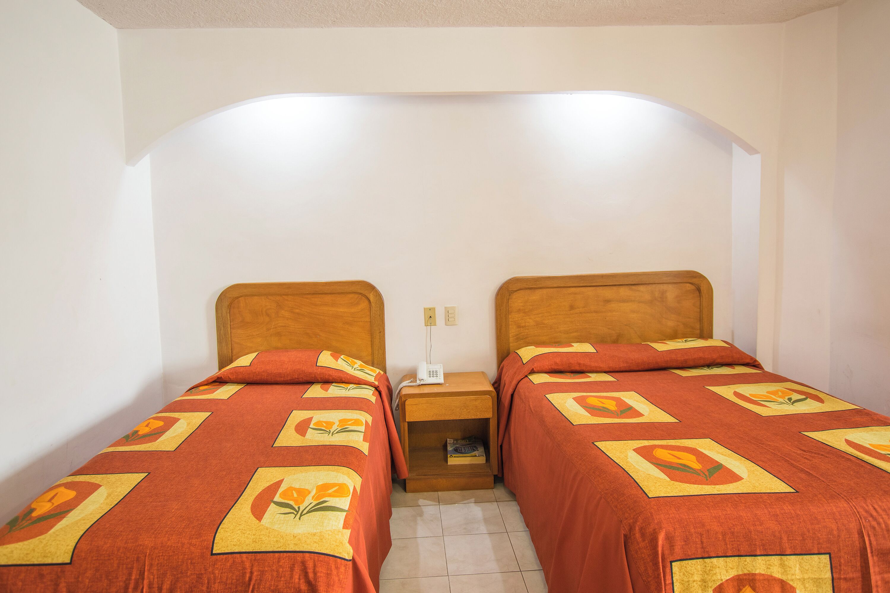 A room with two single beds, each with a wooden headboard and a red bedspread with a floral pattern. There is a small wooden nightstand between the beds with a lamp and a book.