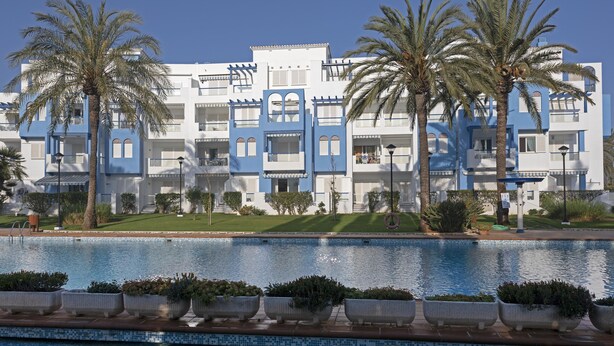 A modern hotel with a pool, palm trees, and a clear blue sky.