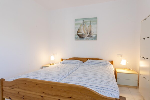 Un dormitorio con cama de madera, una mesita de noche con lámpara, un cuadro colgado en la pared de un velero y un armario.
