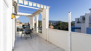 A balcony with a white railing, a table with chairs, and a view of buildings and mountains in the background.