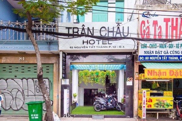 Ein Hotel Eingang mit einem Motorrad davor, einer grünen Mülltonne und einem Schild für einen Imbiss in der Nähe.