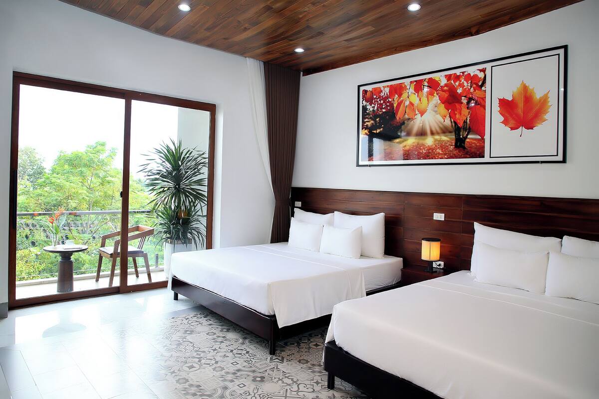 Habitación de hotel con dos camas, un ventanal con vistas a la vegetación, techo de madera y un cuadro de hojas otoñales.