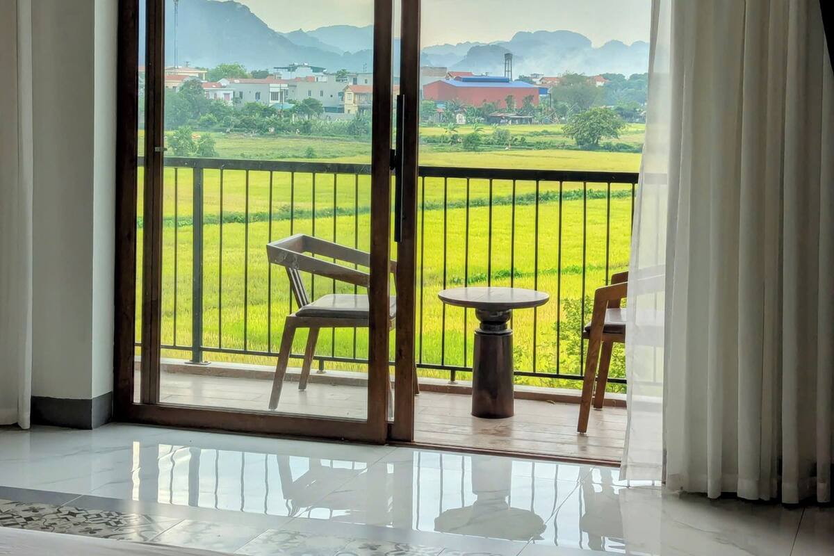Habitación de hotel con un amplio ventanal corredero que da a un balcón con vistas a un campo verde y montañas lejanas.
