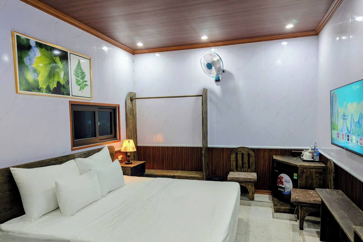 Habitación de hotel con una cama grande, un banco de madera, una mesita con lámpara, un televisor y un ventilador en la pared.