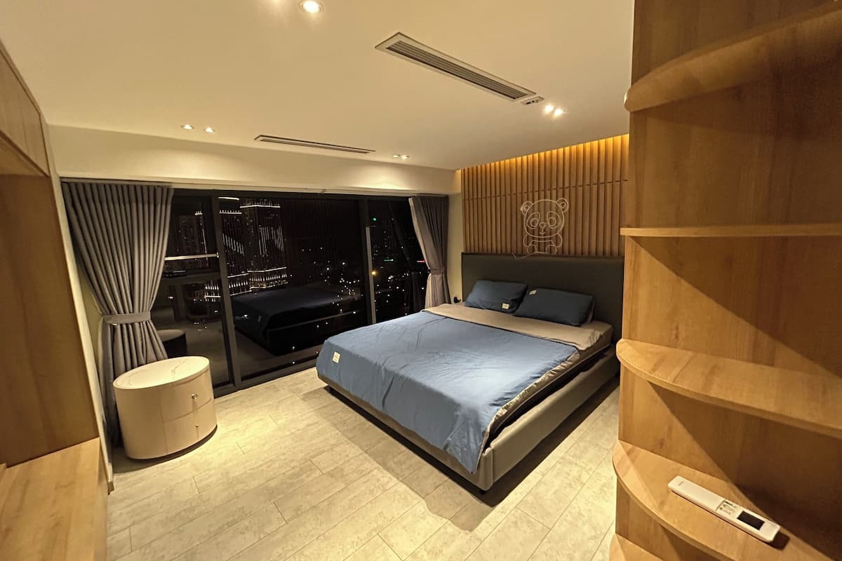 A modern bedroom with a large bed, a bedside table, a built-in wardrobe, and a city view through floor-to-ceiling windows.