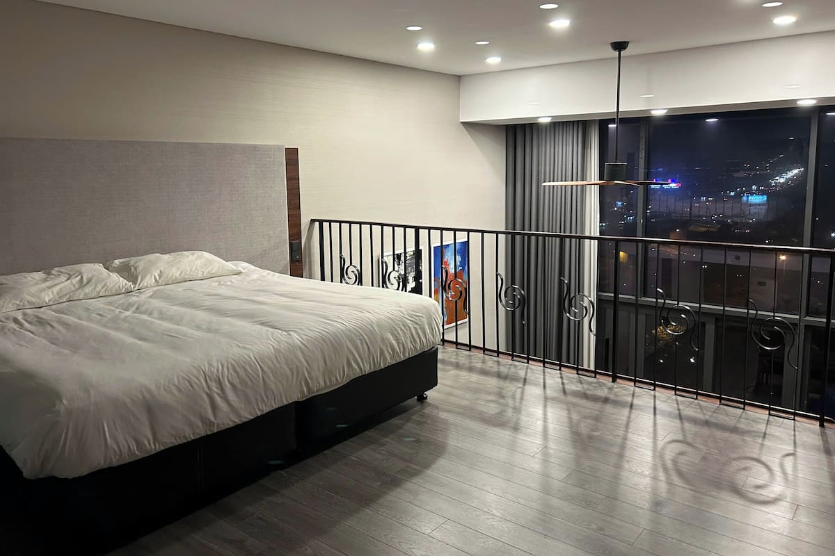 A modern bedroom with a large bed, a balcony with a view of the city at night, and a ceiling with recessed lighting.