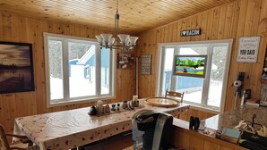 A cozy cabin kitchen with wooden walls, a dining table with a floral tablecloth, a coffee maker, and a view of a snowy landscape through the windows. - Family-Friendly Cottage with all the Amenities You Need!  (Burk's Falls)