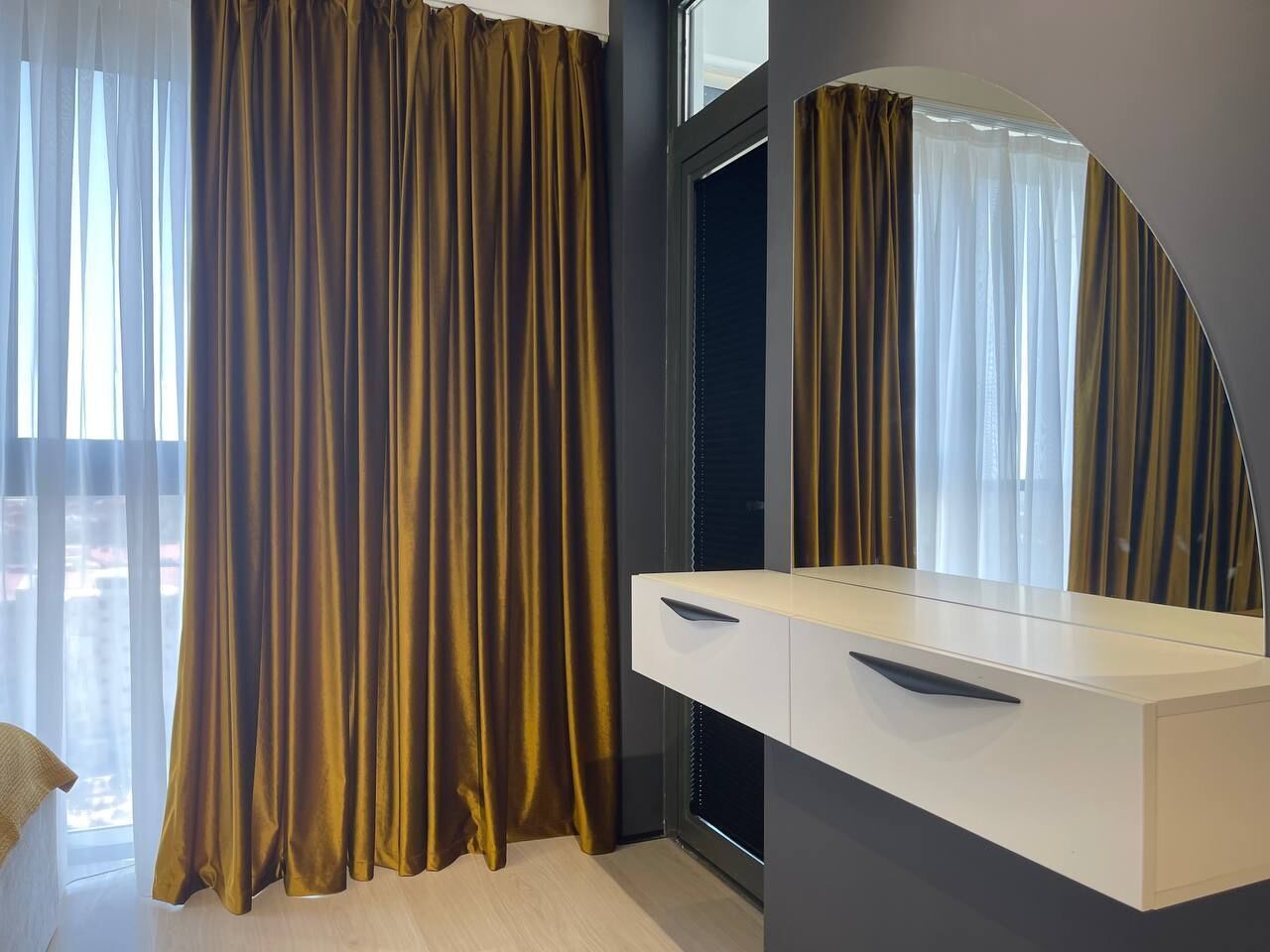 A modern bedroom with a large window, a vanity with a mirror, and a pair of drawers.