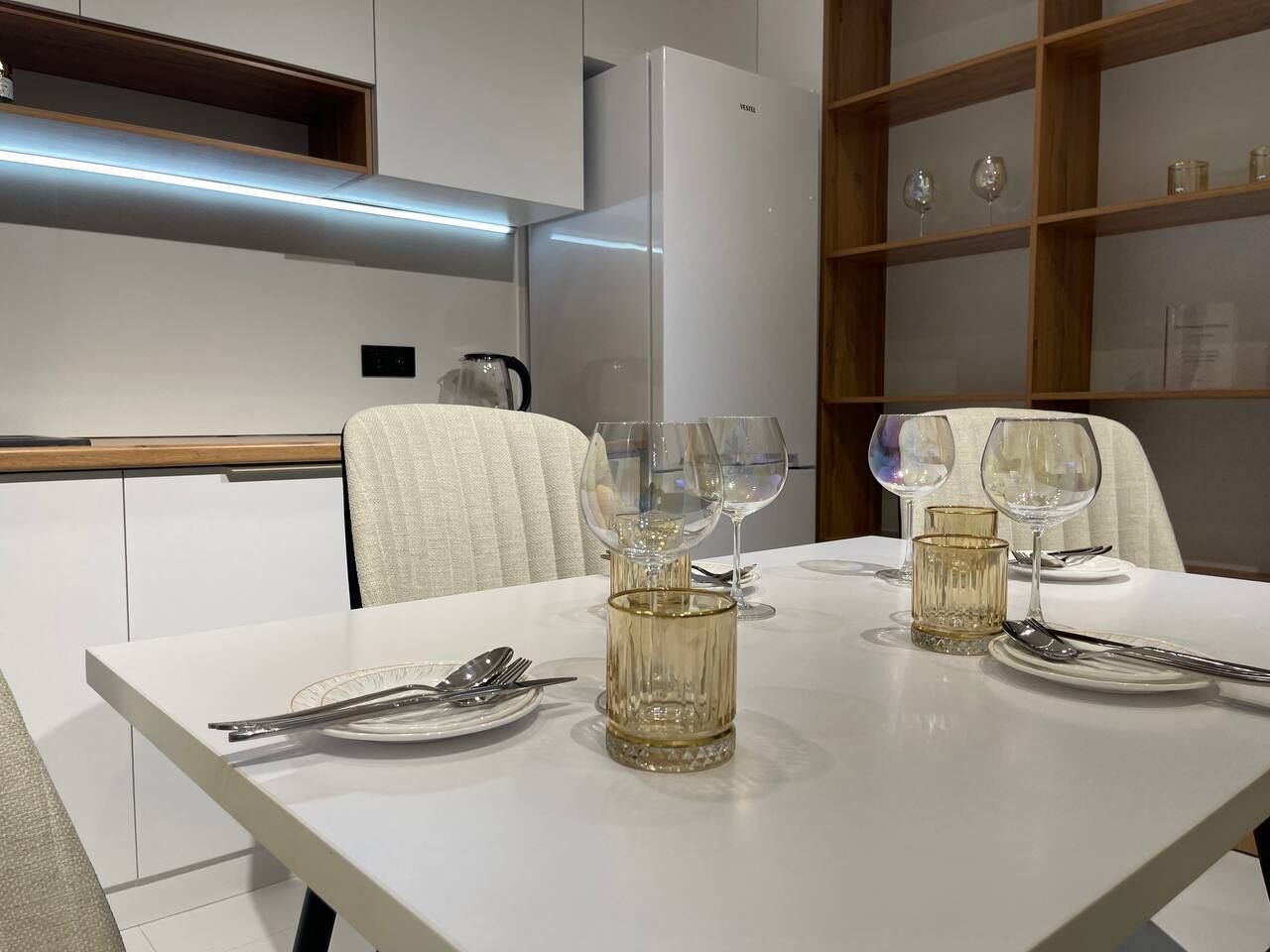 Un espace repas moderne avec une table blanche dressée pour deux personnes, agrémentée de verres et de couverts en verre transparent.