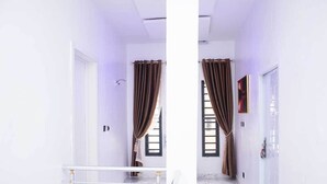 A hallway with white walls, a marble floor, and a white railing. There are brown curtains on the windows and a painting on the wall. - 4-Bedroom Duplex with Private Bathrooms in all Rooms (Lekki-Ajah Express Way)