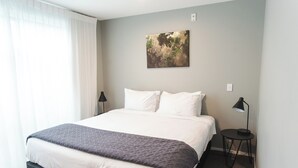 A neatly made bed with white linens and a gray blanket, a small bedside table with a lamp, and a wall-mounted painting.