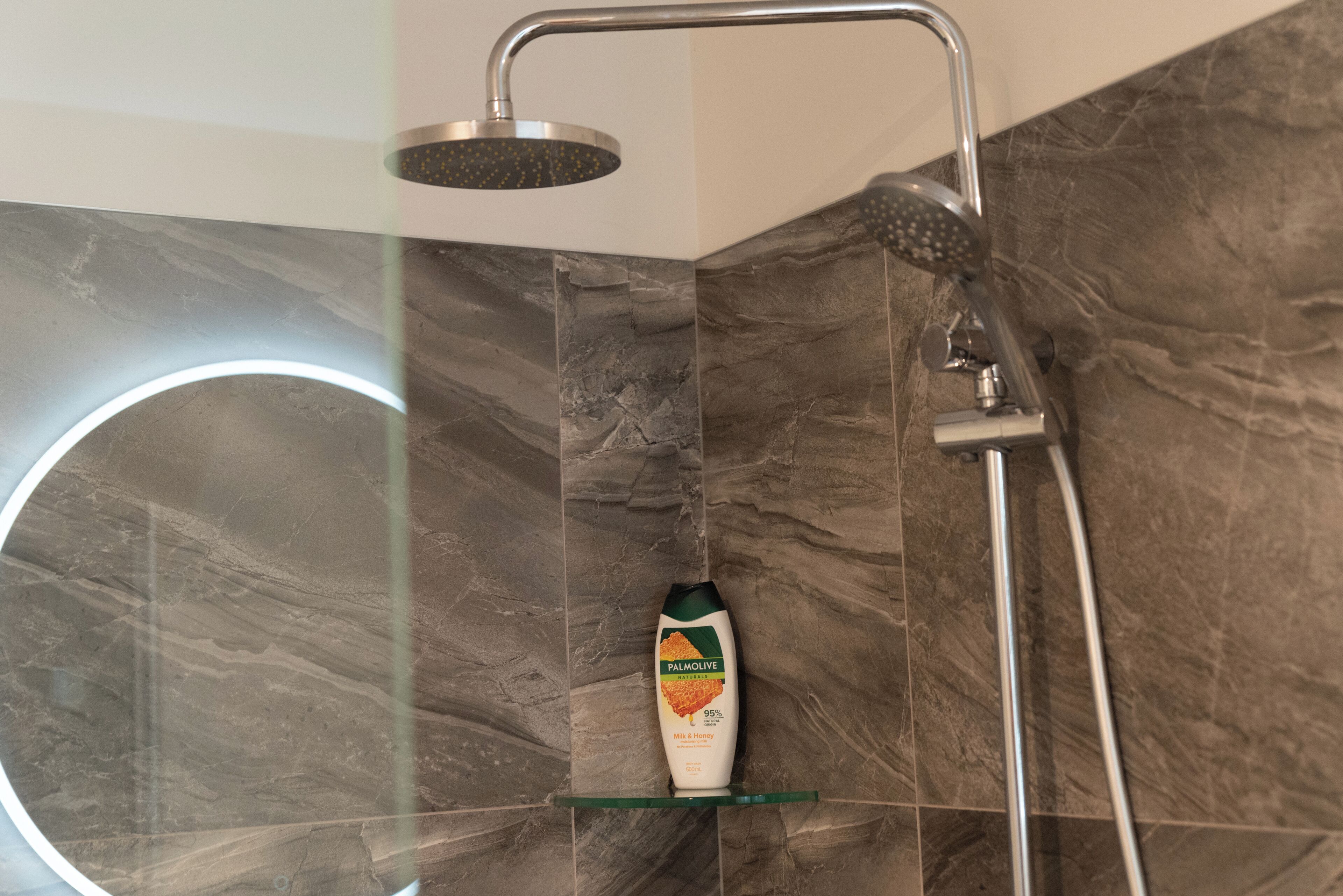 Une douche moderne équipée d’un pommeau de douche à main et d’un robinet mural. Un flacon de shampoing Palmolive est posé sur une étagère en verre.