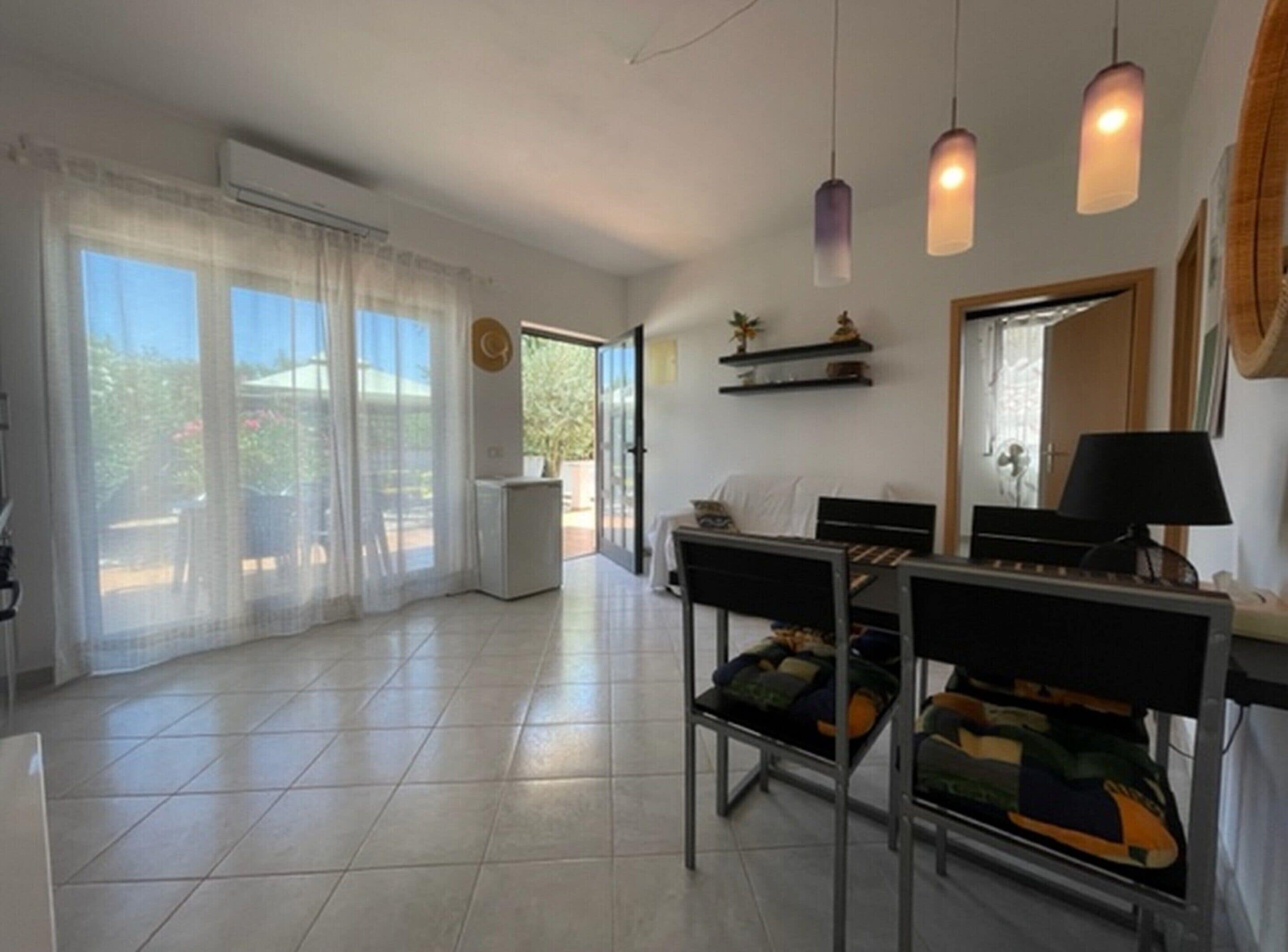 Amplia sala de estar con una mesa de comedor, sillas y un sofá. Hay lámparas colgantes, un televisor y una pequeña mesa con adornos.