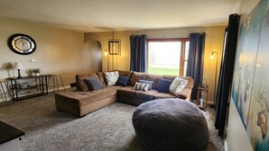 Ein Wohnzimmer mit einer Eckcouch, einem runden Couchtisch, einer Lampe, einer Uhr und Blick ins Freie durch ein Fenster.
