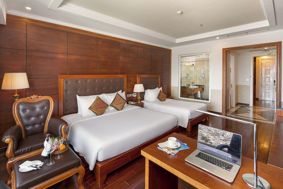 A hotel room with two beds, a desk with a laptop, a chair, and a small table with a cup and saucer.