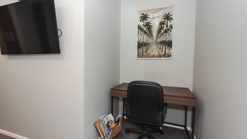 Une pièce comprenant un bureau, un fauteuil de bureau noir, un téléviseur fixé au mur et un tableau représentant des palmiers.