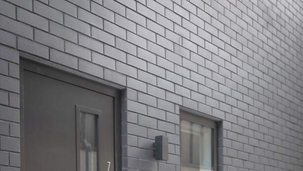 En moderne bygning med en grå murstensfacade, en mørkegrå dør med glasrude og et lille vindue med hvid ramme.