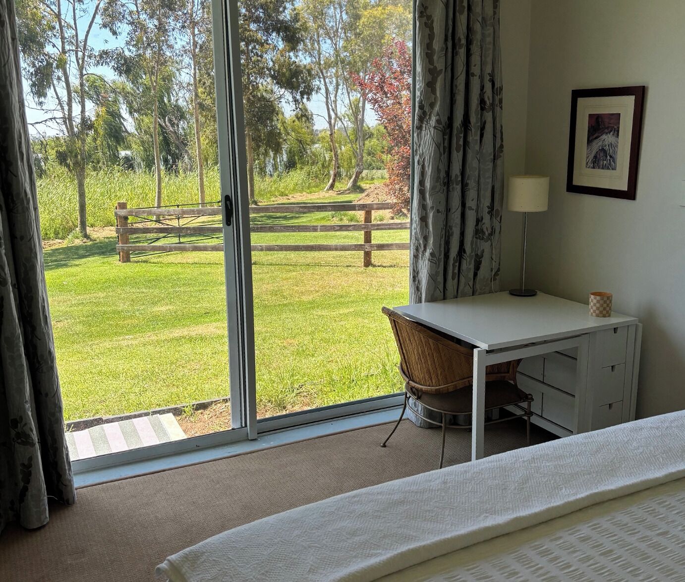 A bedroom with a large sliding glass door leading to a grassy outdoor area, a white desk with a chair, a lamp, and a framed picture on the wall.