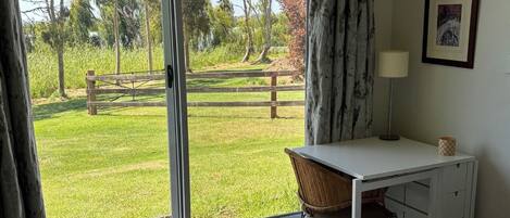 A bedroom with a large sliding glass door leading to a grassy outdoor area, a white desk with a chair, a lamp, and a framed picture on the wall.
