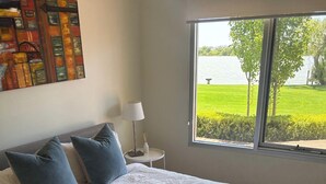 A bedroom with a large bed, two pillows, a bedside table with a lamp, and a window with a view of a green landscape and water. - On The Bend- Absolute Waterfront Retreat (Tailem Bend)