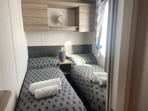 Two single beds with geometric-patterned bedding, a bedside table and lamp, and a shelf above.