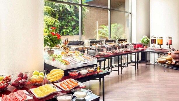 A buffet setup in a spacious room with large windows, offering a view of greenery outside. - Horison Arcadia Wahid Hasyim (Jakarta)
