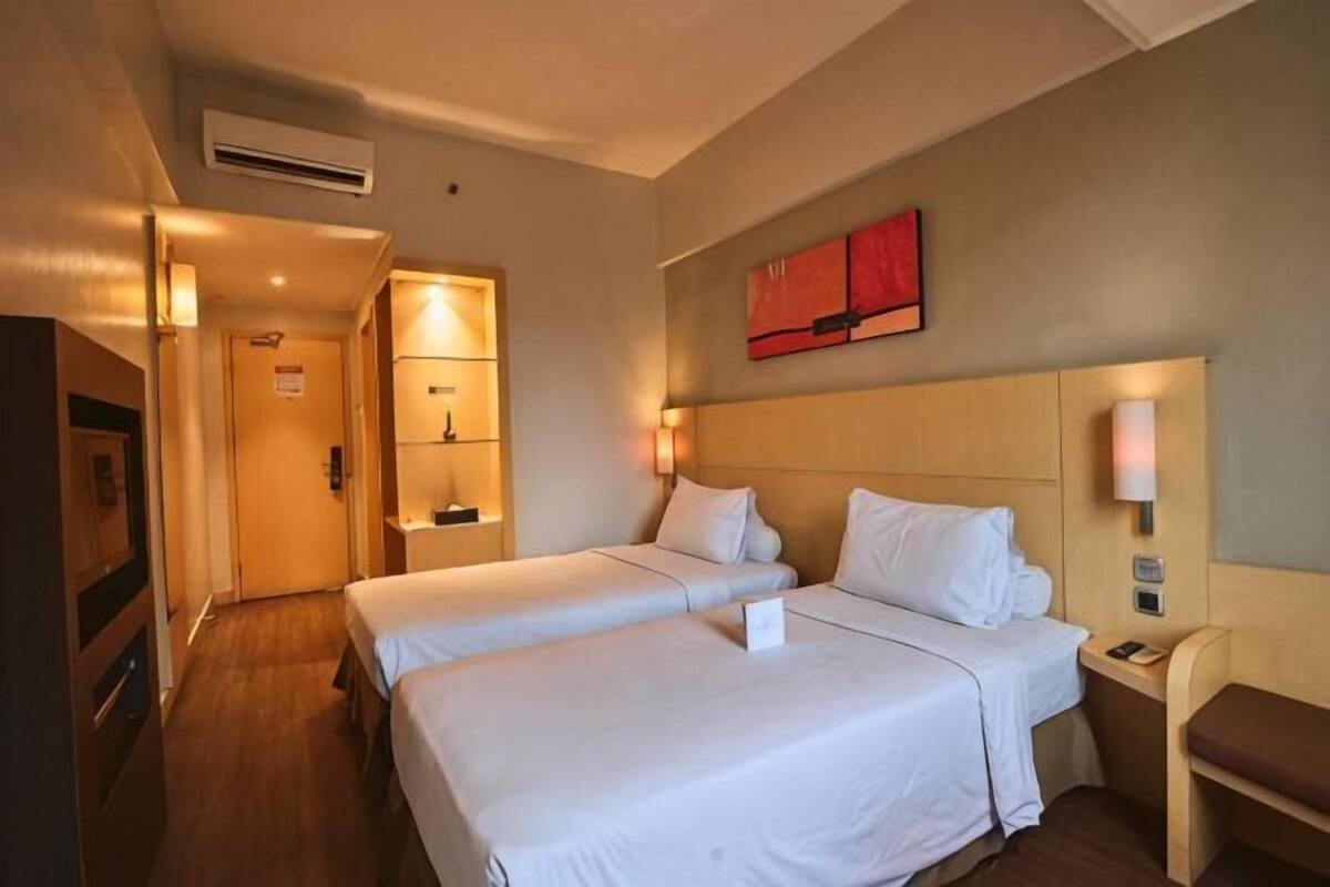 A hotel room with two beds, a desk, a chair, a TV, and a red artwork on the wall.