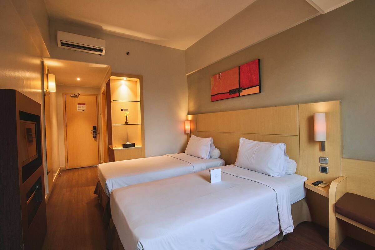 A hotel room with two beds, a wooden headboard, a nightstand with a lamp, and a wall-mounted artwork.