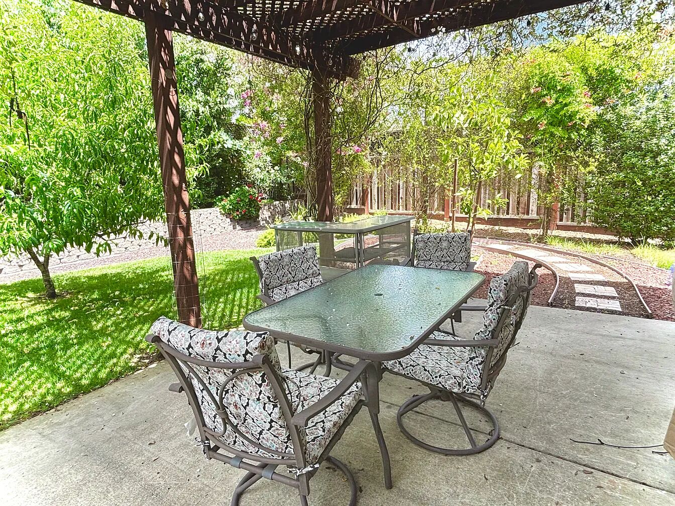 Um pátio externo com mesa de vidro e cadeiras estampadas, cercado por vegetação e árvores.