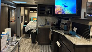 A compact RV interior with a kitchenette, a bed, and a large TV screen displaying an underwater scene. - RV in Art District and Close to the BEACH! Pool & Hot-Tub & Fenced/Pet-Friendly! (Melbourne)