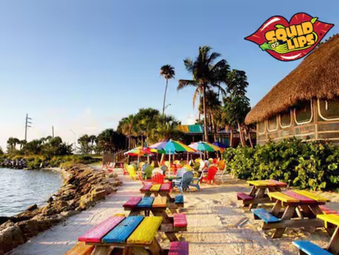 A beachside setting with colorful picnic tables and umbrellas, a thatched-roof building, and palm trees in the background.