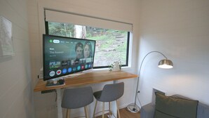 Un salon moderne avec un grand téléviseur à écran plat fixé à un mur blanc, un bureau en bois avec deux chaises, une lampe sur pied et une vue sur un jardin par une fenêtre.