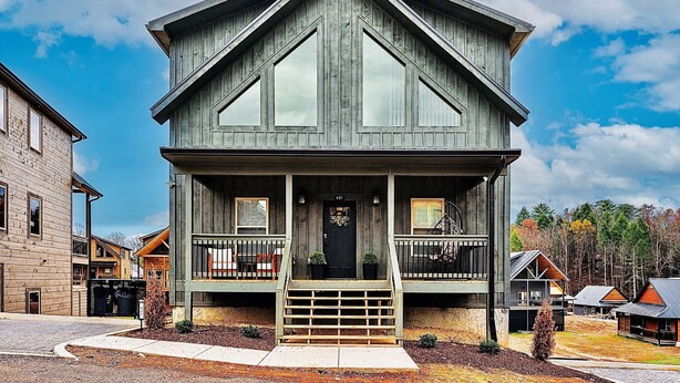 Une maison en bois à deux étages, dotée d’un porche à l’avant, d’une porte d’entrée noire et d’un petit jardin devant.