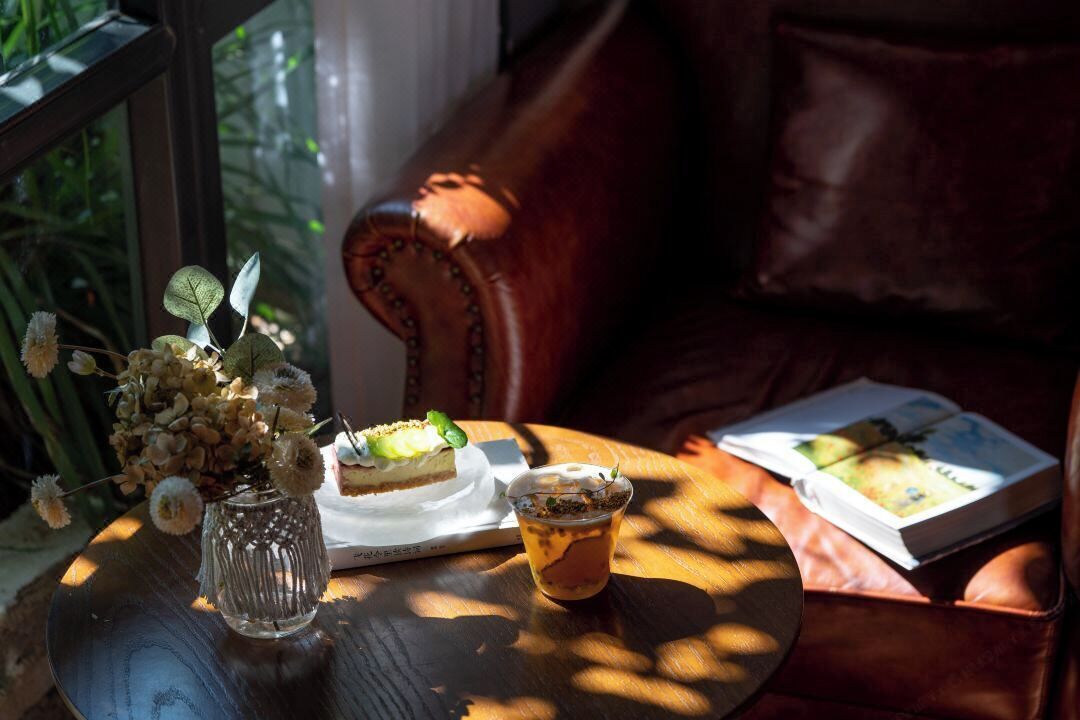 A table with a glass vase, a dessert, and a drink, with a book and a leather couch in the background.