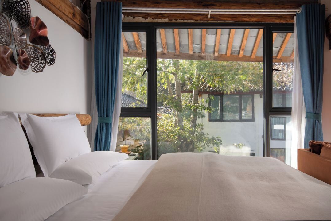 A bedroom with a large bed, wooden ceiling beams, and a view of greenery outside.