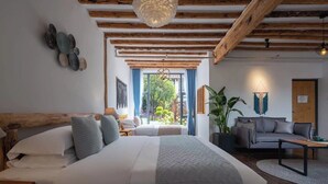 Ein Schlafzimmer mit einem großen Bett, Holzbalken, einer Pendelleuchte, Blick ins Grüne und einem grauen Sofa im Wohnbereich.