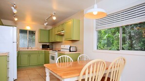 A compact kitchen with green cabinets, a wooden dining table, and white chairs. There is a window with blinds, a refrigerator, and a stove. - Yonegan Maui Getaways - Haiku - Unit 2 (Haiku-Pauwela)