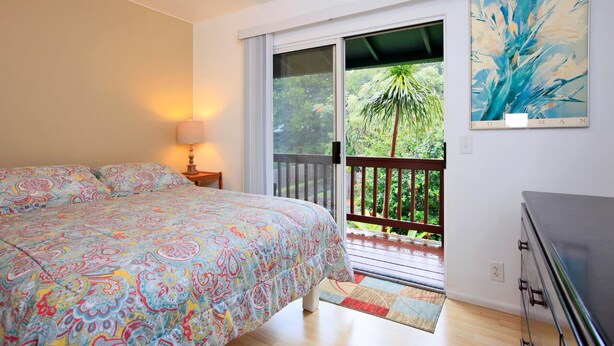 A bedroom with a bed, a nightstand, a lamp, a painting, and a balcony with a view of greenery. - Yonegan Maui Getaways - Haiku - Unit 2 (Haiku-Pauwela)