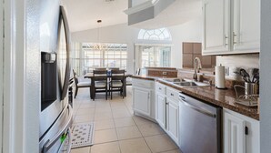 A modern kitchen with white cabinets, granite countertops, and stainless steel appliances. There is a dining area with a table and chairs, and a large window providing natural light.