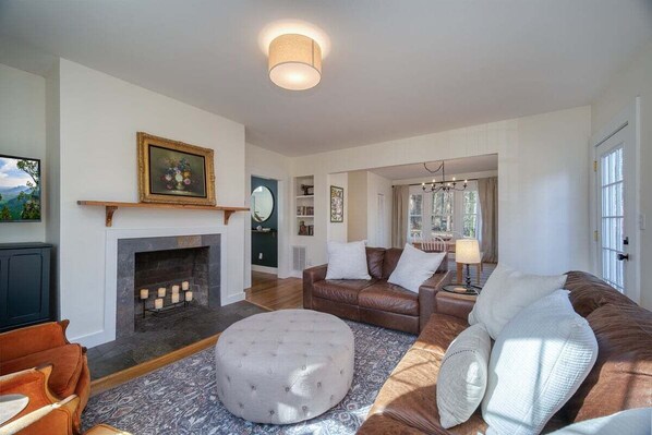 A living room with a fireplace, leather sofas, a round ottoman, and a chandelier. - Monthly Rental 5 Minutes to Downtown! (Brevard)
