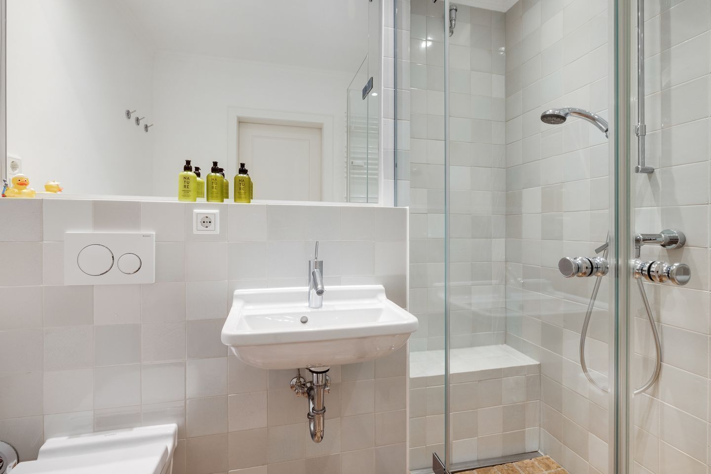 A modern bathroom with a white sink, glass shower enclosure, and a shelf with toiletries.