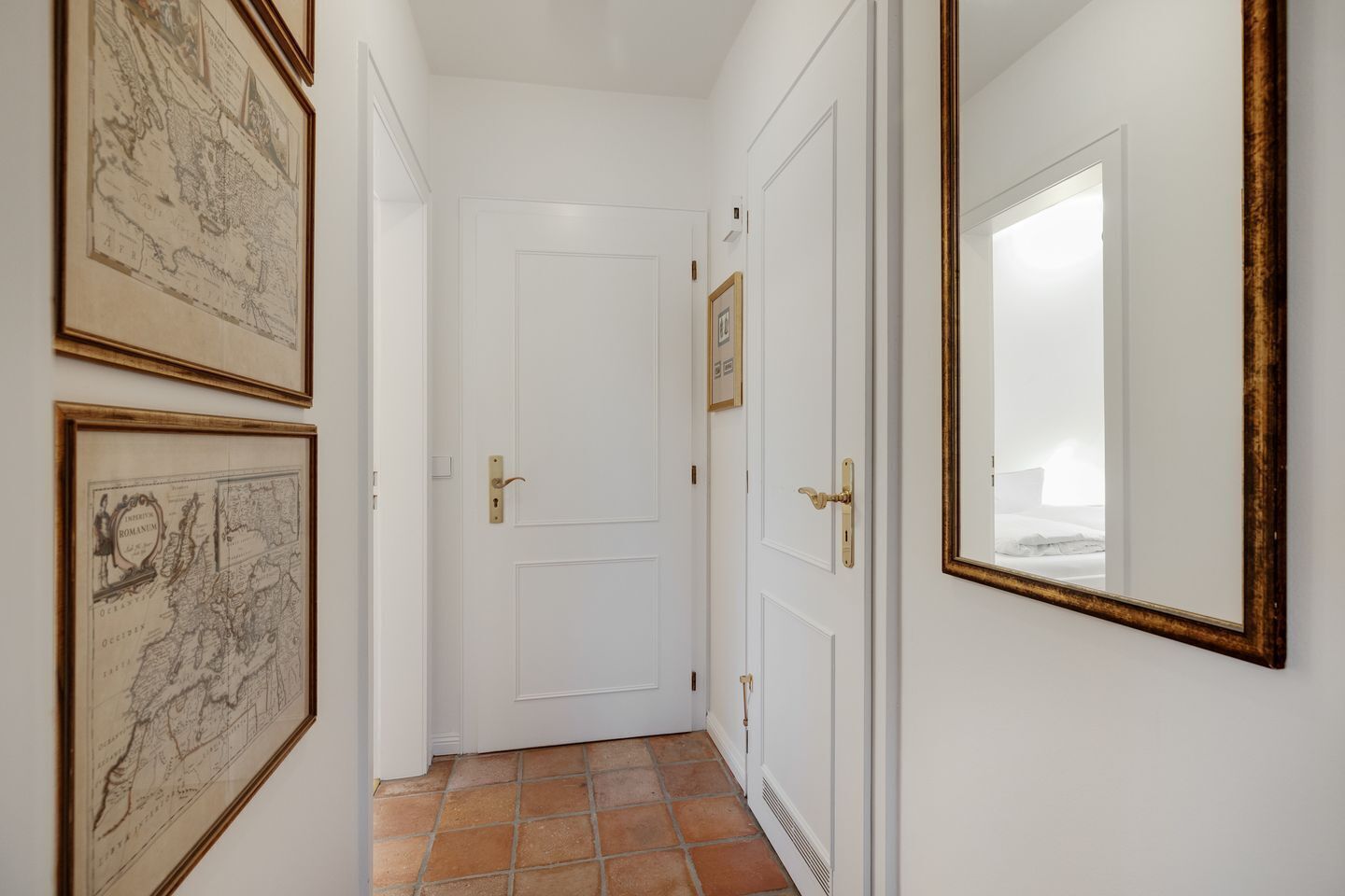 A hallway with a white door, framed maps on the wall, and a mirror reflecting a bed.