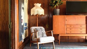 Een houten kast met lades, een stoel met gestreepte kussens, een vloerlamp en een poster aan de muur.