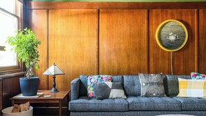 Een gezellige woonkamer met een houten lambrisering, een blauwe bank met kleurrijke kussens, een potplant en een lamp op een bijzettafeltje.