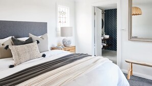 A neatly made bed with a grey headboard, a bedside table with a lamp, and a view into a room with a herringbone-patterned wall.
